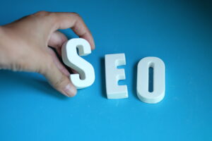 “Hand arranging white 3D letters spelling SEO on a blue background, representing search engine optimization and website ranking strategies.”